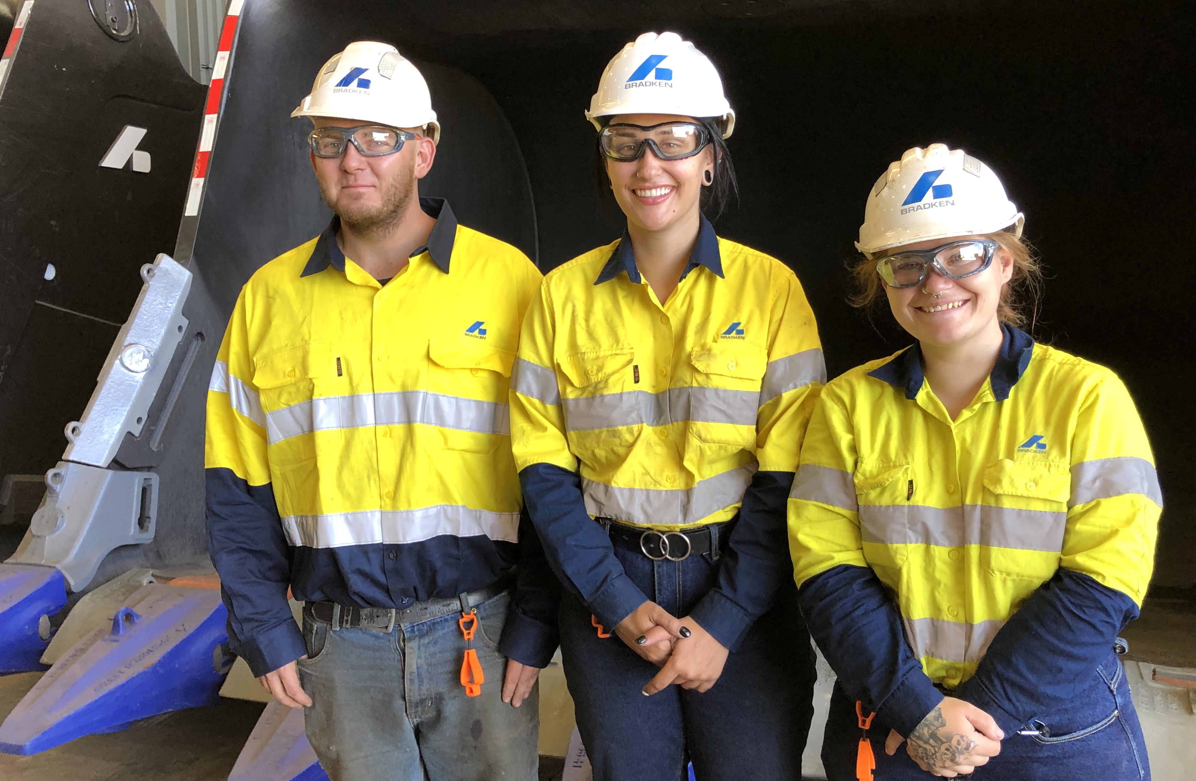Three people, two women and one man, stand in front of an excavator bucket wearing yellow hi-viz uniforms and white hard hats with the Bradken logo on them.
