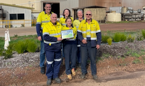 A group of men and women wearing yellow hi-viz uniforms stand outside a foundry site. Immediately behind them is a garden.