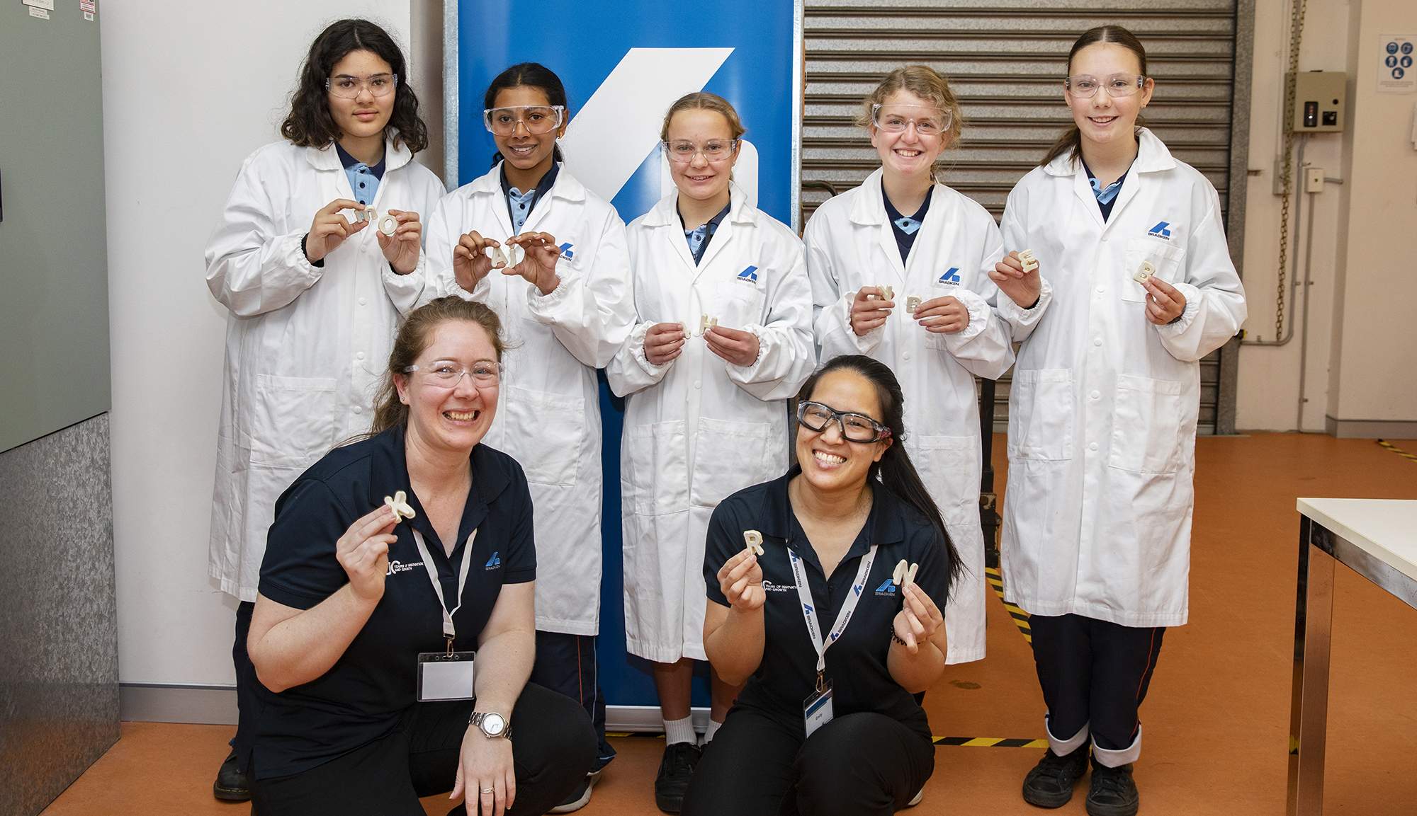 A group of school girls in white lab coats stand behind two crouching women wearing Bradken branded polo shirts. All hold small plaster cast letters.